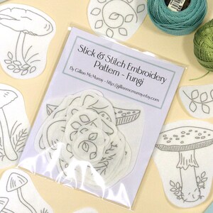 May include: A Stick & Stitch Embroidery Pattern - Fungi kit featuring a clear plastic packet of white adhesive embroidery designs, several loose mushroom and leaf pattern cutouts, and two round skeins of teal and olive green embroidery thread.
