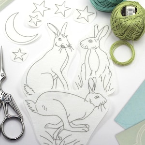 May include: A flat lay of embroidery supplies. The image shows white fabric cutouts of rabbits, stars, and a crescent moon. Two spools of thread, one teal and one green, and a pair of decorative scissors are also present.