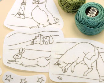 May include: A set of white fabric patches with black line art illustrations of a puffin with a lighthouse, a rabbit with a building, a fox, a crescent moon, and three stars, accompanied by two round balls of green and teal embroidery thread.