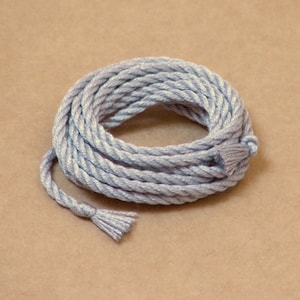 May include: Coiled light blue rope with a tassel end. The rope is tightly wound, creating a circular shape. The tassel is made of the same material and color as the rope. The background is a solid tan color.