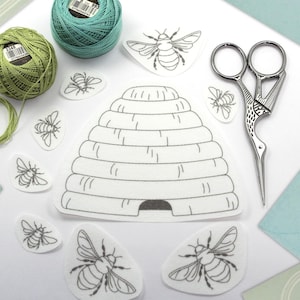 May include: A craft kit with a bee theme, featuring green and teal thread spools, bee-shaped felt cutouts, a beehive cutout, and decorative scissors. The items are arranged on a white surface with light blue and green accents.