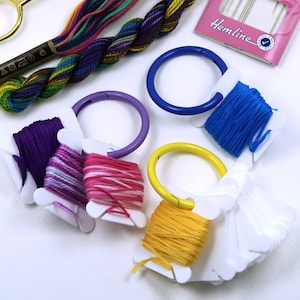 Puede incluir: Un conjunto de suministros de bordado, que incluye tijeras doradas, hilo de bordar colorido y un paquete de agujas de bordado. También se ven varios carretes de hilo en varios colores, como morado, rosa, azul y amarillo.