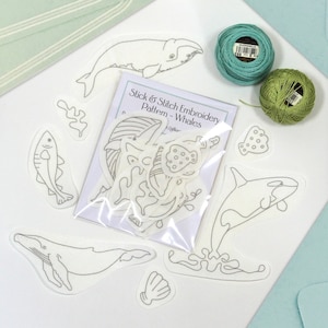May include: A set of white embroidery patterns featuring whales, fish, and seaweed. The patterns are packaged in a clear plastic bag with a white card that reads "Stick & Stitch Embroidery Pattern - Whales". Two spools of embroidery floss are included, one teal and one green.