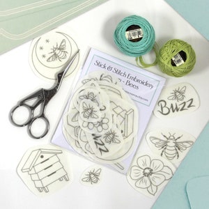 May include: A set of white felt bee-themed embroidery patterns featuring a beehive, bees, flowers, and the word "Buzz". The set includes a pair of scissors and two spools of embroidery floss, one green and one blue.