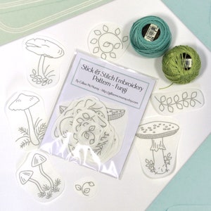 22 Fungi Stick & Stitch embroidery patterns on self adhesive water soluble stabilizer sitting beside green threads and envelopes.