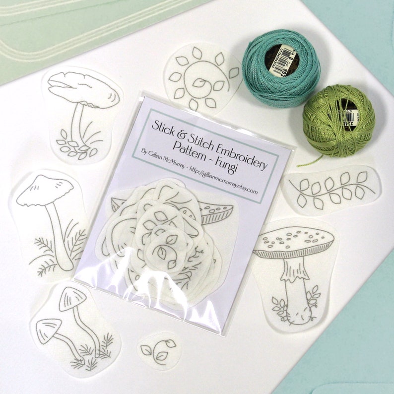 22 Fungi Stick & Stitch embroidery patterns on self adhesive water soluble stabilizer sitting beside green threads and envelopes.