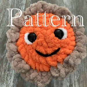 May include: A crocheted orange sun with a smiling face and black eyes. The sun is surrounded by a gray crocheted halo.
