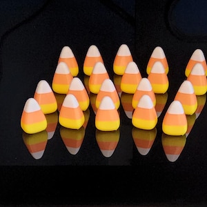 May include: A collection of candy corn pieces arranged on a reflective black surface. Each piece features a classic tri-color design: white tip, orange center, and yellow base. The candies are evenly spaced, creating a visually appealing pattern.