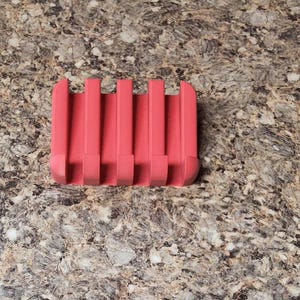 May include: A coral-colored, rectangular kitchen utensil with five vertical slots. The item is made of a solid material and has a curved base. It is designed to hold multiple items. The object is on a speckled, brown surface.