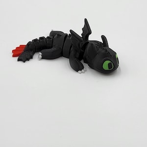 May include: A black dragon figurine with green eyes and red tail fins. The dragon is lying down on a white surface. The figurine has a detailed design, including wings and articulated joints.
