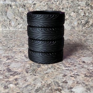 May include: A stack of four black rubber tires with a detailed tread pattern. The tires are cylindrical and appear to be for a small-scale vehicle or model. The tires are stacked on top of each other.
