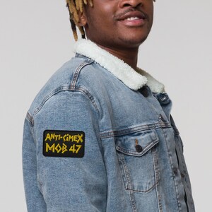Peut inclure: Une veste en jean bleue avec un patch brodé noir et jaune qui dit "ANTI-CIMEX MOB 47".