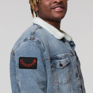 May include: A blue denim jacket with a black patch on the left sleeve. The patch has red embroidered text that reads "DISCLOSE".