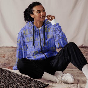 Puede incluir: Una sudadera con capucha azul con un patrón amarillo y negro de jeroglíficos. La modelo lleva la sudadera con pantalones negros y zapatillas blancas.