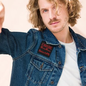 Peut inclure: Une personne portant une veste en jean bleue avec un patch noir qui dit "CRUDE" en lettres rouges. Le patch dit également "Extreme Guitars" et "DISCARD" en lettres rouges plus petites.