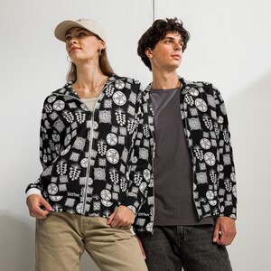 May include: Black zip-up hoodie and a black button-up shirt, both with a repeating white pattern of geometric shapes and symbols. The hoodie has a white zipper and cuffs. The person on the left is wearing a tan baseball cap and khaki pants.