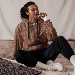 May include: A brown, orange, and teal patterned zip-up hoodie with a black zipper. The model is wearing the hoodie with black pants and white sneakers.