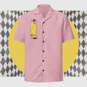以下が含まれることがあります： キャンプカラーの半袖ピンクシャツ。 黄色のコートと帽子を着た人のグラフィックが特徴です。 黒いボタンと襟の近くに小さなラベルが付いています。