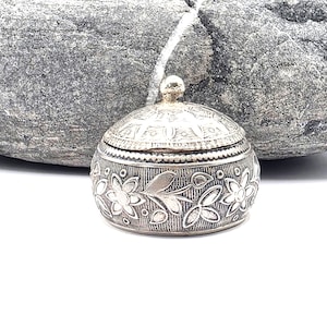 Könnte beinhalten: Eine kleine, runde Schmuckdose aus silberfarbenem Metall mit einem komplizierten, geprägten Blumenmuster an den Seiten und einem passenden gewölbten Deckel mit einem kleinen kugelförmigen Knauf. Die Oberfläche hat ein dunkles, oxidiertes Finish.