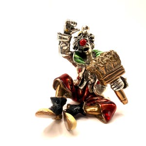 Könnte beinhalten: Detaillierte Figur eines Clowns in sitzender Position. Der Clown trägt eine rote Hose, eine silberne Jacke und schwarze Stiefel mit beigefarbenen Sohlen. Der Clown hat eine rote Nase, einen grünen Kragen und hält einen Pinsel.