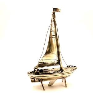 Puede incluir: Figura de velero plateado con mástil, vela y bandera. El barco tiene un casco detallado y está sostenido por pequeñas patas. El velero es un objeto decorativo.