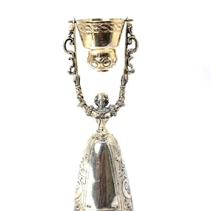 Op de afbeelding: Een zilverkleurige metalen beker met een figuur met opgeheven armen die een kleinere, afneembare kom boven het hoofd ondersteunt. De basis heeft de vorm van een brede, conische rok met gegraveerde patronen en de kom heeft een textuur aan de rand.