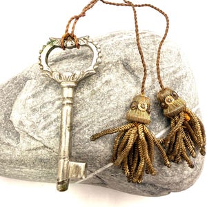 Puede incluir: Una llave plateada antigua con un asa ornamentada descansa sobre una piedra gris. Dos borlas doradas decorativas cuelgan de un cordón marrón. La llave tiene un diseño vintage detallado, lo que sugiere un artículo decorativo o de colección.