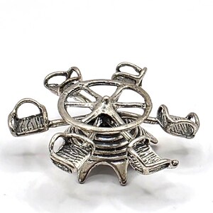 Puede incluir: Un dije de metal plateado con forma de carrusel de feria en miniatura. El objeto presenta un marco circular central con cinco asientos pequeños e individuales unidos por brazos delgados, todo en un acabado metálico con detalles visibles.