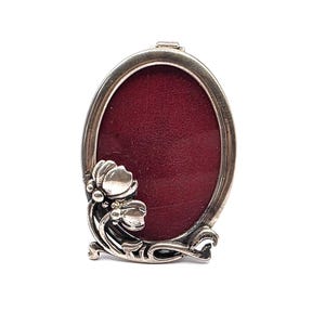 Könnte beinhalten: Ein ovaler Bilderrahmen aus Silber mit einer tiefroten Samtrückseite. Der Rahmen ist an der unteren linken Ecke mit einem metallischen Blumenmotiv verziert, das aus zwei abgerundeten Blütenknospen und geschwungenen Stielen besteht.