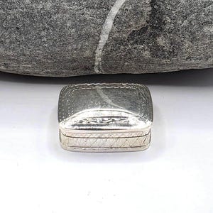 Pode incluir: Uma pequena caixa retangular de metal prateado com tampa articulada. A tampa apresenta uma borda gravada com padrões de pontos e linhas, enquanto as laterais da base são decoradas com um design repetitivo de hachuras diagonais cruzadas.