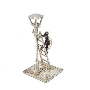 May include: A silver-toned miniature sculpture of a person climbing a ladder to a street lamp. The lamp has a square top and is mounted on a textured base. The figure is detailed, with visible features. The sculpture is set against a white background.