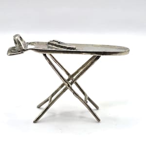 Op de afbeelding: Een miniatuur zilverkleurige metalen strijkplank met een inklapbaar X-vormig onderstel en een klein, plat strijkijzer op het oppervlak. Het object heeft een gladde, metalen afwerking en is afgebeeld in zijaanzicht tegen een effen achtergrond.