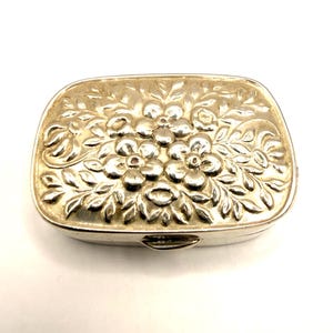 Puede incluir: Una caja rectangular plateada con un diseño floral en la tapa. La caja tiene un cierre de bisagra y probablemente está hecha de metal. El patrón floral es en relieve y detallado.