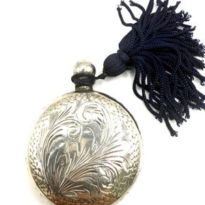 Peut inclure: Une flasque ronde argentée avec un motif floral orné. La flasque a un gland bleu foncé attaché au sommet. La flasque est probablement en métal et a une finition brillante.