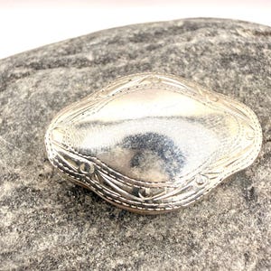 Puede incluir: Un broche de metal plateado con un centro liso y abombado y una forma alargada de diamante. Los bordes presentan intrincados patrones lineales grabados y pequeños detalles circulares que enmarcan la superficie pulida y reflectante de la pieza.