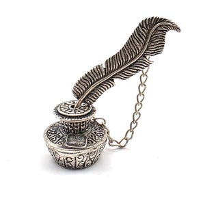 May include: A silver-toned inkwell with a decorative feather quill and chain. The inkwell has intricate patterns and a detailed design. The feather quill is positioned at an angle, and the chain is attached to the inkwell.