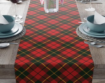 May include: A red, green, and gold plaid table runner. The runner is rectangular and covers the center of a wooden table. The table is set with plates, bowls, silverware, and wine glasses. A vase of pink flowers sits in the center.