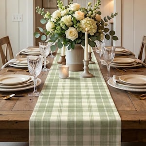 May include: A rustic wooden dining table set for a meal, featuring a green and white plaid table runner. The table is set with plates, silverware, and wine glasses, and a floral centerpiece. A chandelier hangs above.