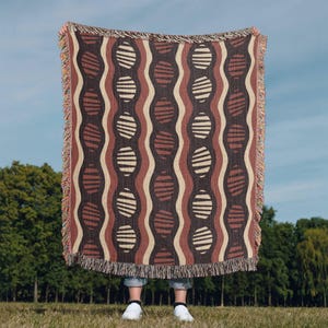 May include: A woven throw blanket with a repeating pattern of brown, beige, and white oval shapes and wavy lines. The blanket has a fringed edge and is held up against a backdrop of green trees and a blue sky.