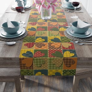 May include: A patchwork table runner with heart motifs in various colours. The runner displays squares with patterns like polka dots and stripes. The table is set with plates, bowls, cutlery, and wine glasses, creating a dining setting.