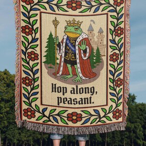 May include: Woven throw blanket featuring a regal frog wearing a crown and holding a scepter, with the text "Hop along, peasant." The design includes a castle, trees, and floral borders in red, green, and blue.