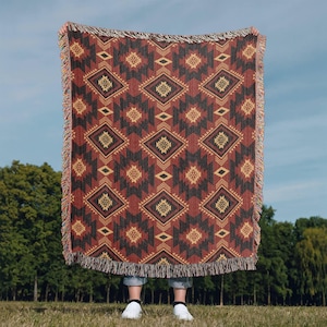 May include: A woven throw blanket with a geometric pattern in shades of red, black, and tan. The blanket features a diamond and zigzag design, with fringed edges. The blanket is held up against a backdrop of green trees and a blue sky.