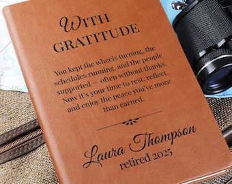 May include: A brown leather journal with the words "WITH GRATITUDE" and a personalized message. The journal is inscribed with "Laura Thompson retired 2025". A camera and map are in the background.