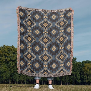 May include: A woven throw blanket with a geometric diamond pattern in shades of blue, brown, and tan. The blanket has a fringed edge and is held up against a backdrop of green trees and a blue sky.