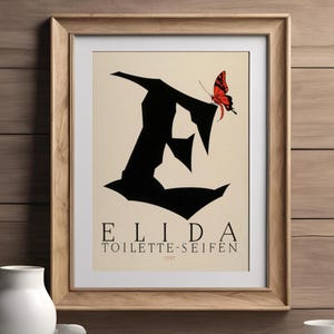 May include: Framed art print featuring a black silhouette of the letter "E" with a red and black butterfly. The print includes the text "ELIDA TOILETTE-SEIFEN" in a serif font, set against a cream background. The artwork is in a wooden frame.