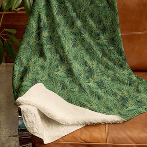 May include: A green throw blanket with a pattern of pine needles is draped over a brown leather couch. The blanket has a soft, cream-colored sherpa lining, providing warmth and comfort. The blanket is a cozy home decor item.