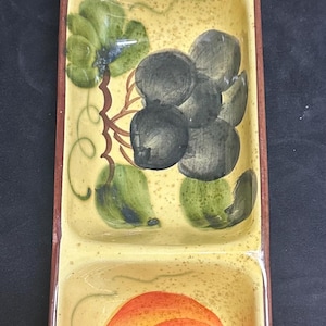 Puede incluir: Una fuente rectangular dividida con fondo amarillo y borde marrón. La fuente está decorada con imágenes pintadas a mano de frutas, incluyendo naranjas, uvas y hojas verdes. La fuente mide aproximadamente 30 cm de largo.