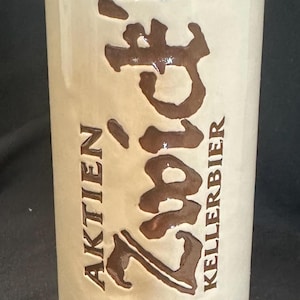 May include: A tall, cylindrical, cream-coloured ceramic beer mug featuring the brown embossed text Zwick'l, Aktien, and Kellerbier. A circular emblem at the base depicts a figure in armour with the text Bayreuther Bierbrauerei AG Aktien Seit 1857.