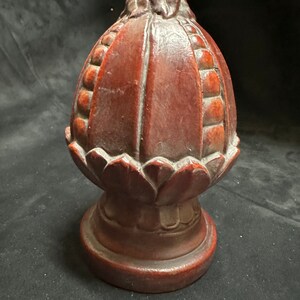 May include: A dark brown wooden finial with a detailed carved design. The finial has a rounded top with a pointed tip and a base that resembles a lotus flower.