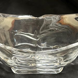 May include: A clear glass decorative bowl with a wavy design. The bowl has a rectangular base and curved sides that extend into looped handles. The glass has a rippled texture, adding visual interest to the piece.